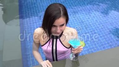 美丽的女孩在游泳池游泳，喝蓝色的鸡尾酒玛格丽塔。